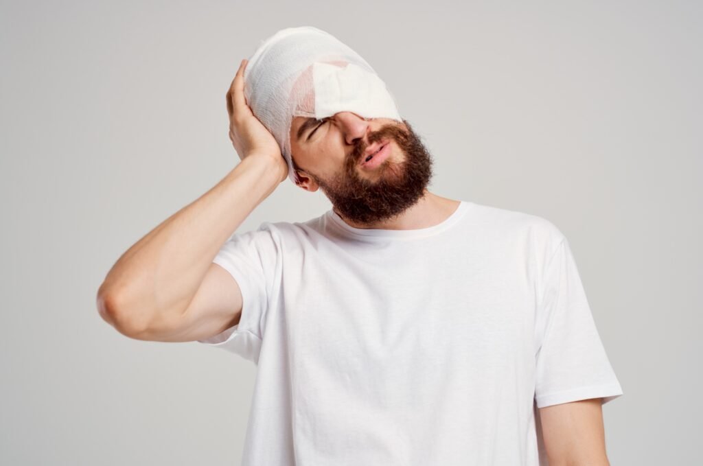 A man with a bandage over his head and eye holding his head in pain.